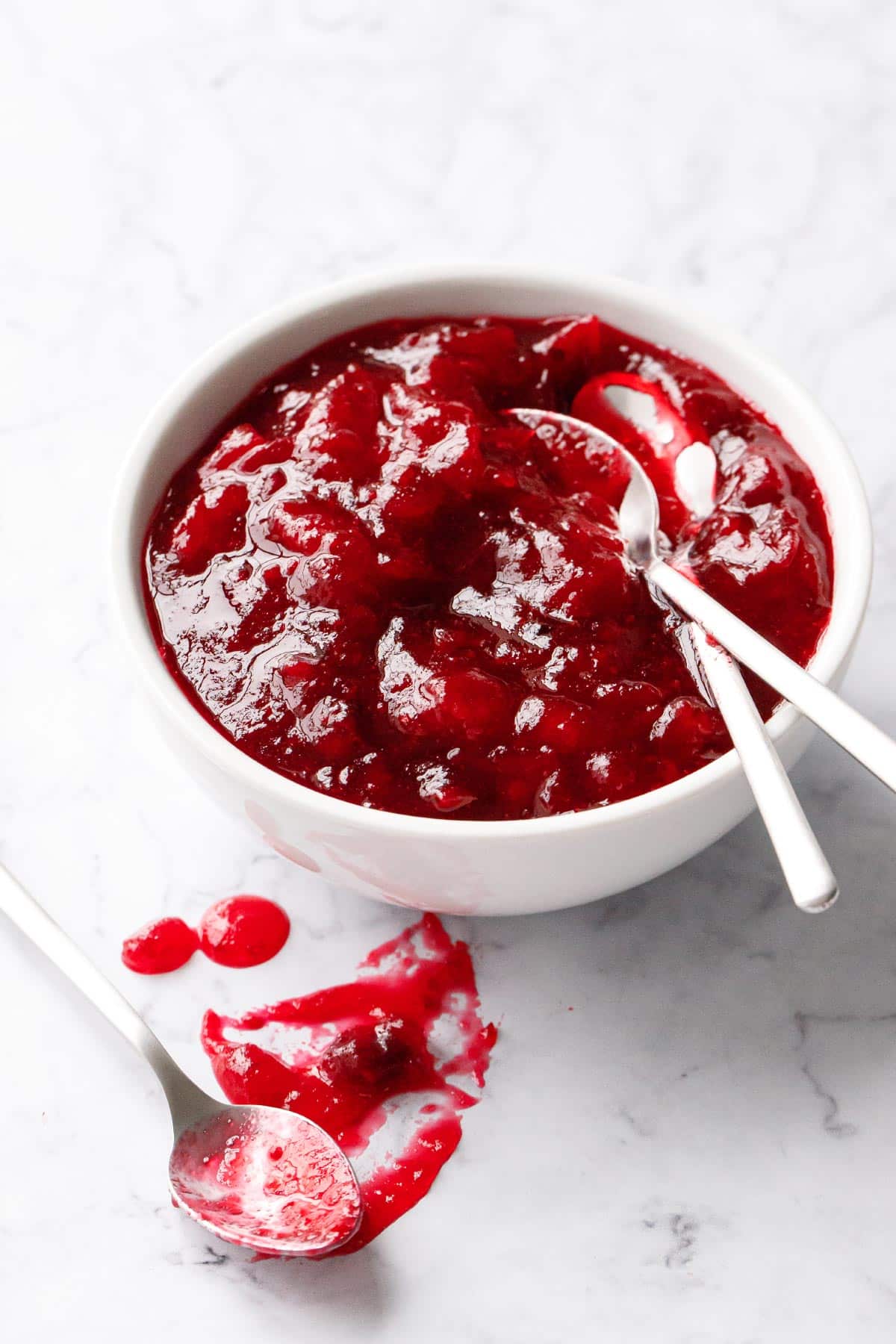 White bowl filled with Classic Cranberry Sauce and two spoons, plus another spoon with a smear of cranberry sauce on the marble surface.