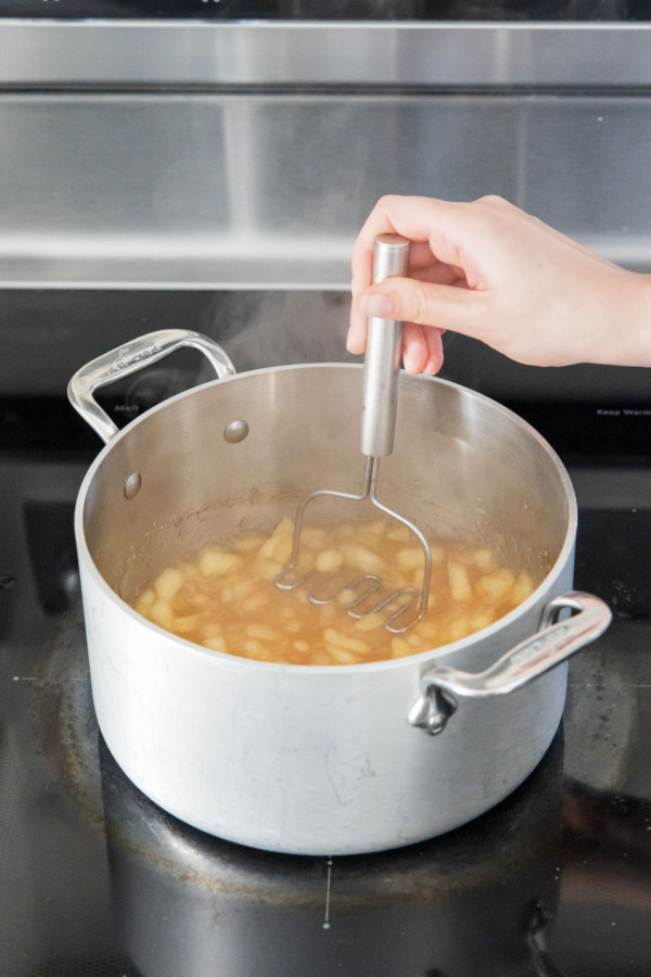 Using a potato masher to smash softened apple pieces into a chunky sauce.