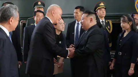 Xinhua Kim shakes hands with Chinese politician Cai Qi on the train platform, surrounded by officials and uniformed guards. His daughter stands behind him, wearing a black dress suit.