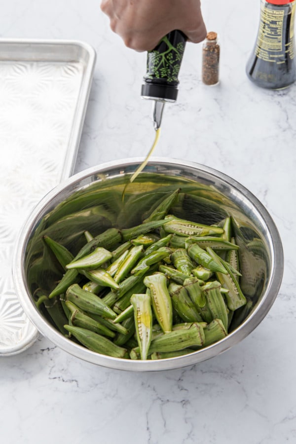 Drizzling halved okra pods with olive oil in a stainless mixing bowl.