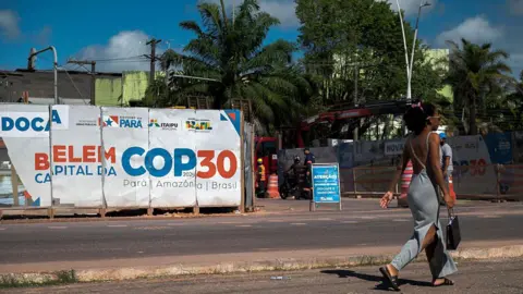 AFP via Getty Images A woman walks past an infrastructure project under way for COP30 in Belem. She has her hair up and is wearing a grey dress and carrying a black bag. Misaligned posters in white, blue and orange read "Belem Capital Da COP30".