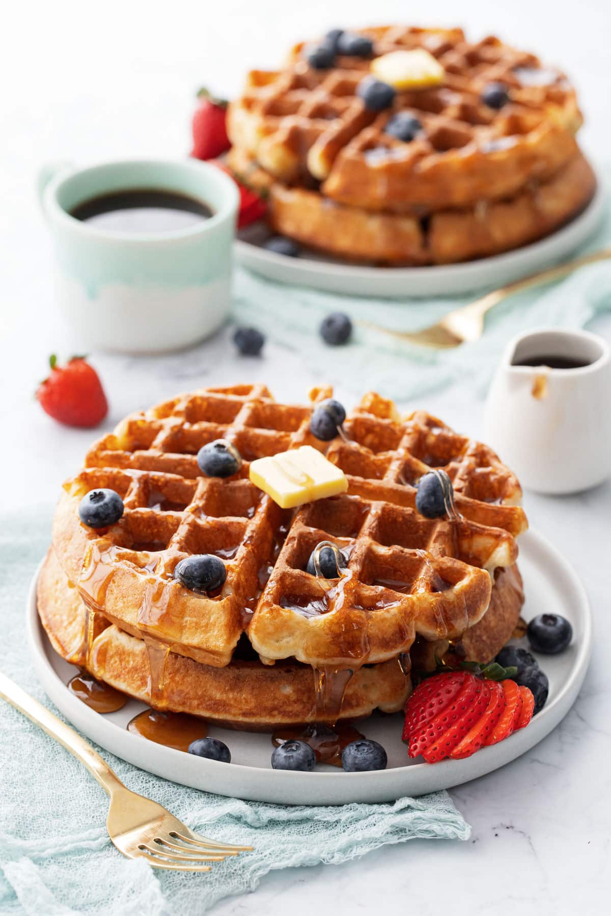 Plates with stacks of golden brown sourdough Belgian waffles topped with berries, a pat of butter and drizzled syrup.