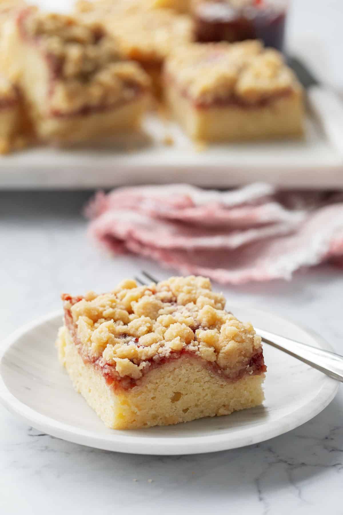 Square of Jammy Sour Cream Crumb Cake on a marble dessert plate with fork, napkin and the rest of the cut cake in the background.