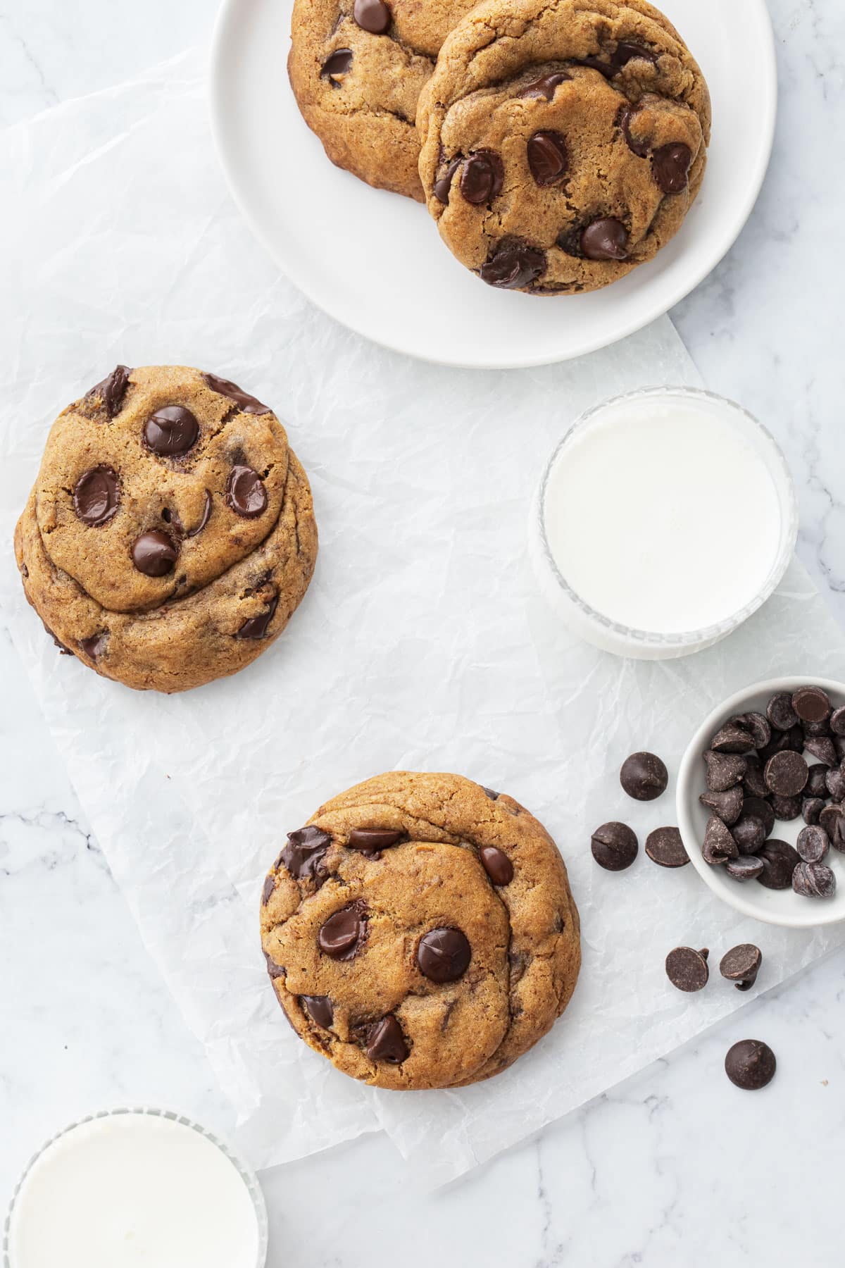 Overhead, extra large Brown Butter Chocolate Chip Cookies on white marble with parchment paper and a glass of milk.