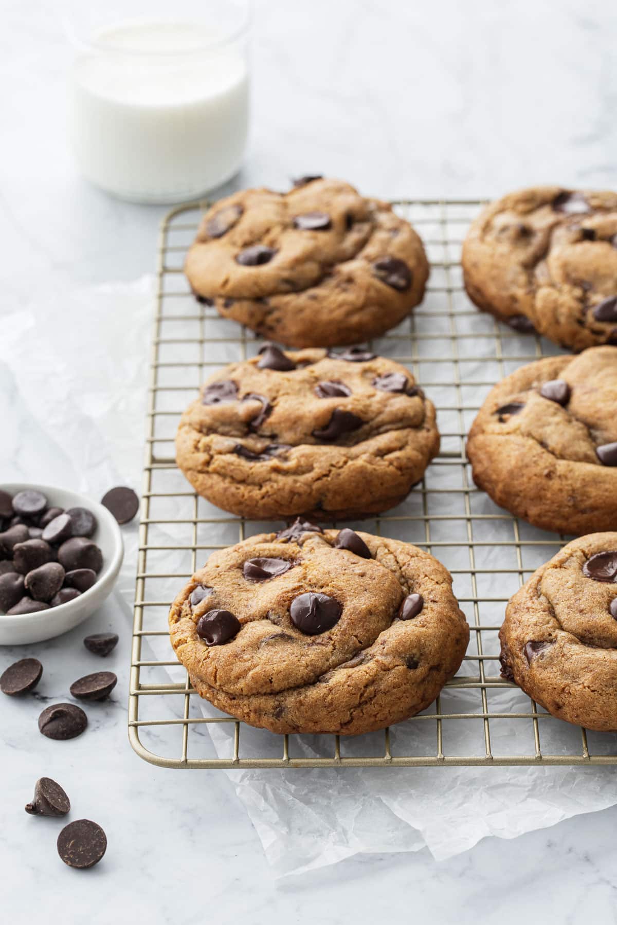 Six extra-large Brown Butter Chocolate Chip Cookies on a wire rack with a glass of milk and bowl of chocolate chips on the side.