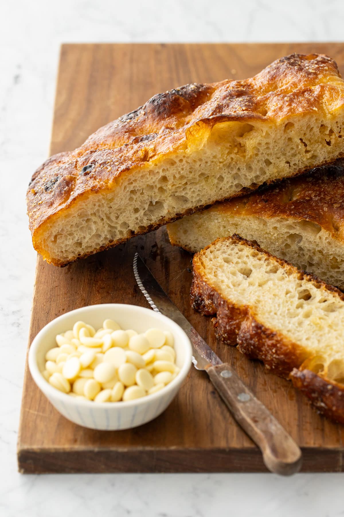 Slices of White Chocolate Focaccia Bread on a wooden cutting board with a bowl of white chocolate pieces and a serrated bread knife on the side.