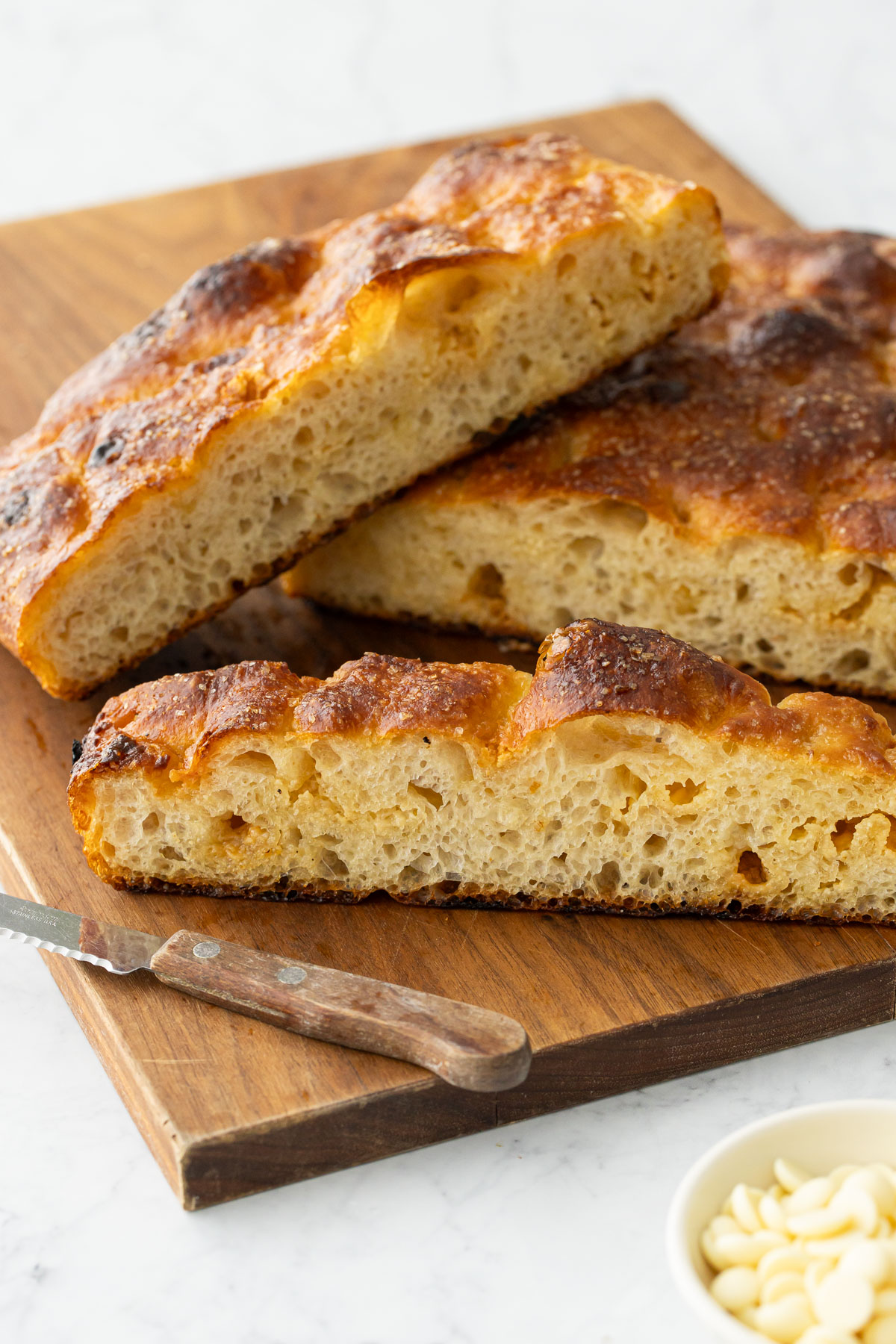 Slices of White Chocolate Focaccia Bread arranged on a wooden cutting board with a serrated knife.
