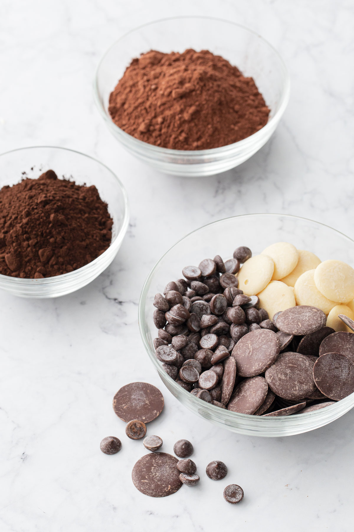 Bowls filled with various kinds of chocolate and cocoa powder on a white marble background.