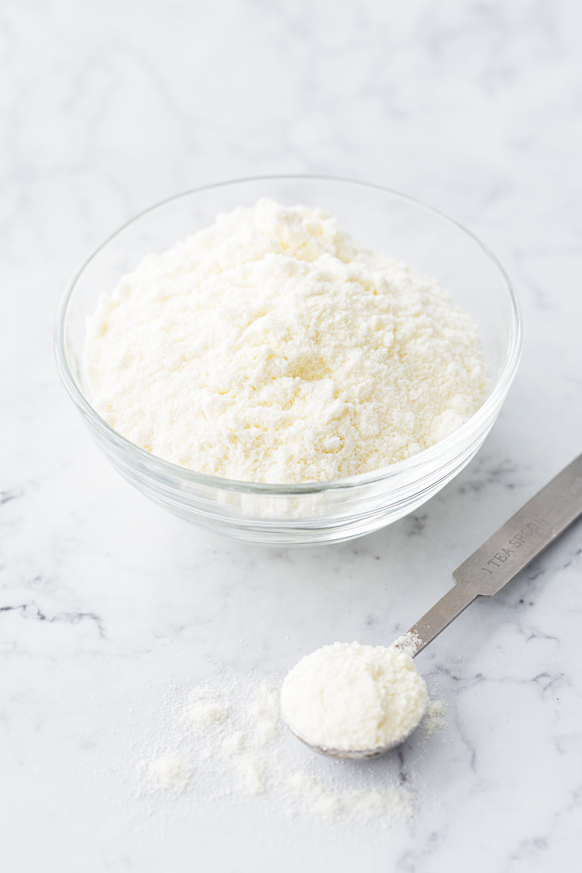 Clear bowl with dry milk powder, with a teaspoon of milk powder messily arranged on the side.