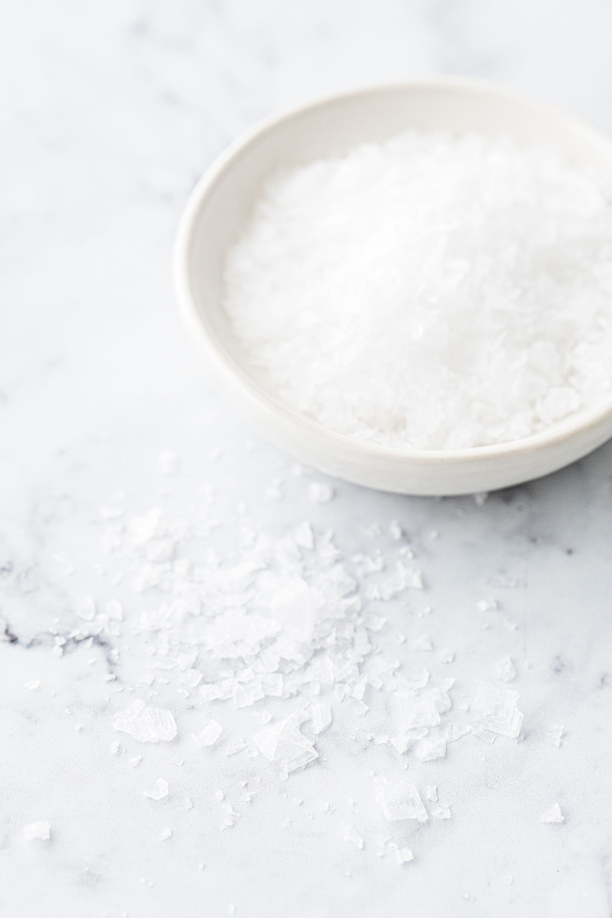 Flake sea salt in a white bowl, some crystals spilled onto the side to show the large crystal structure.