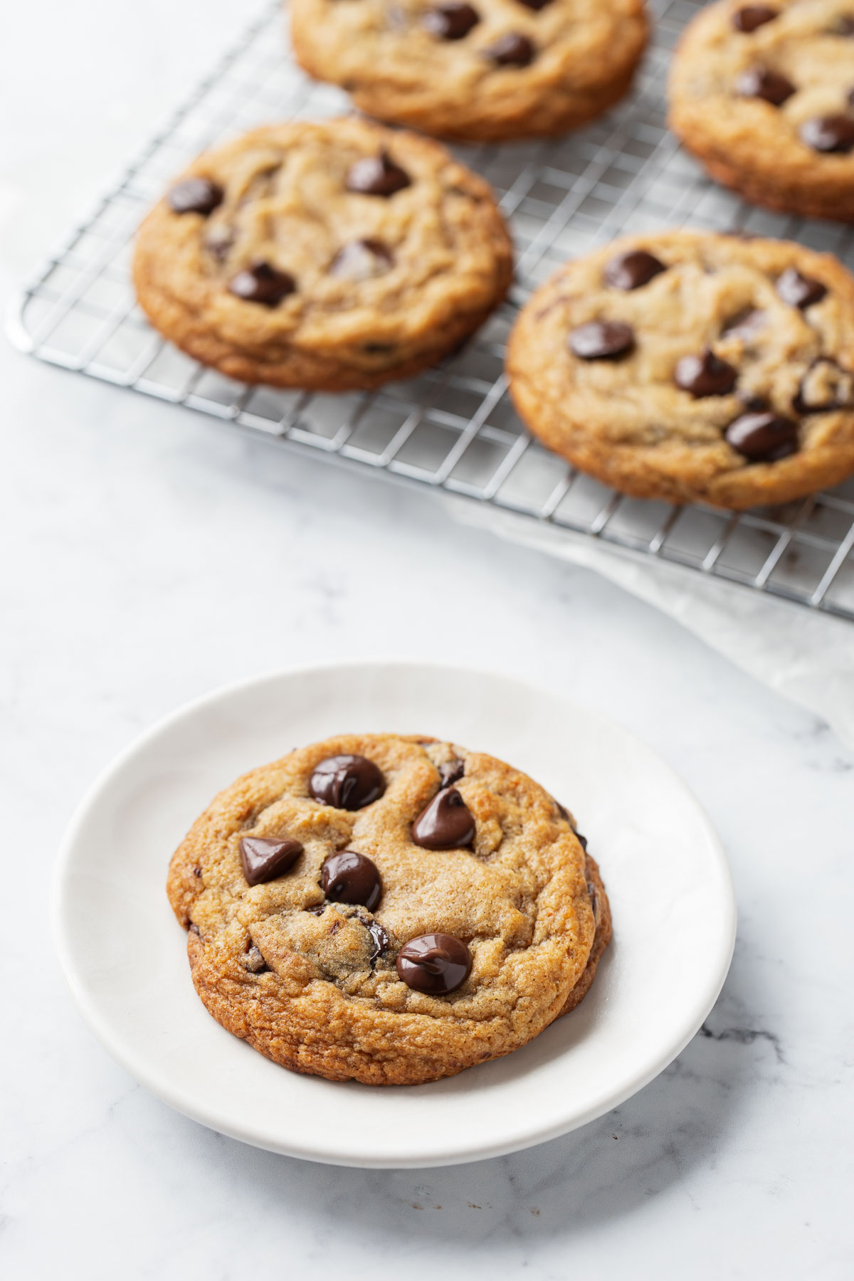 One Brown Butter Banana Bread Chocolate Chip Cookie on a white plate, with a wire rack of more cookies in the background.