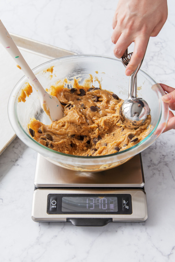 A large cookie scoop spooning cookie dough from mixing bowl on top of a kitchen scale.