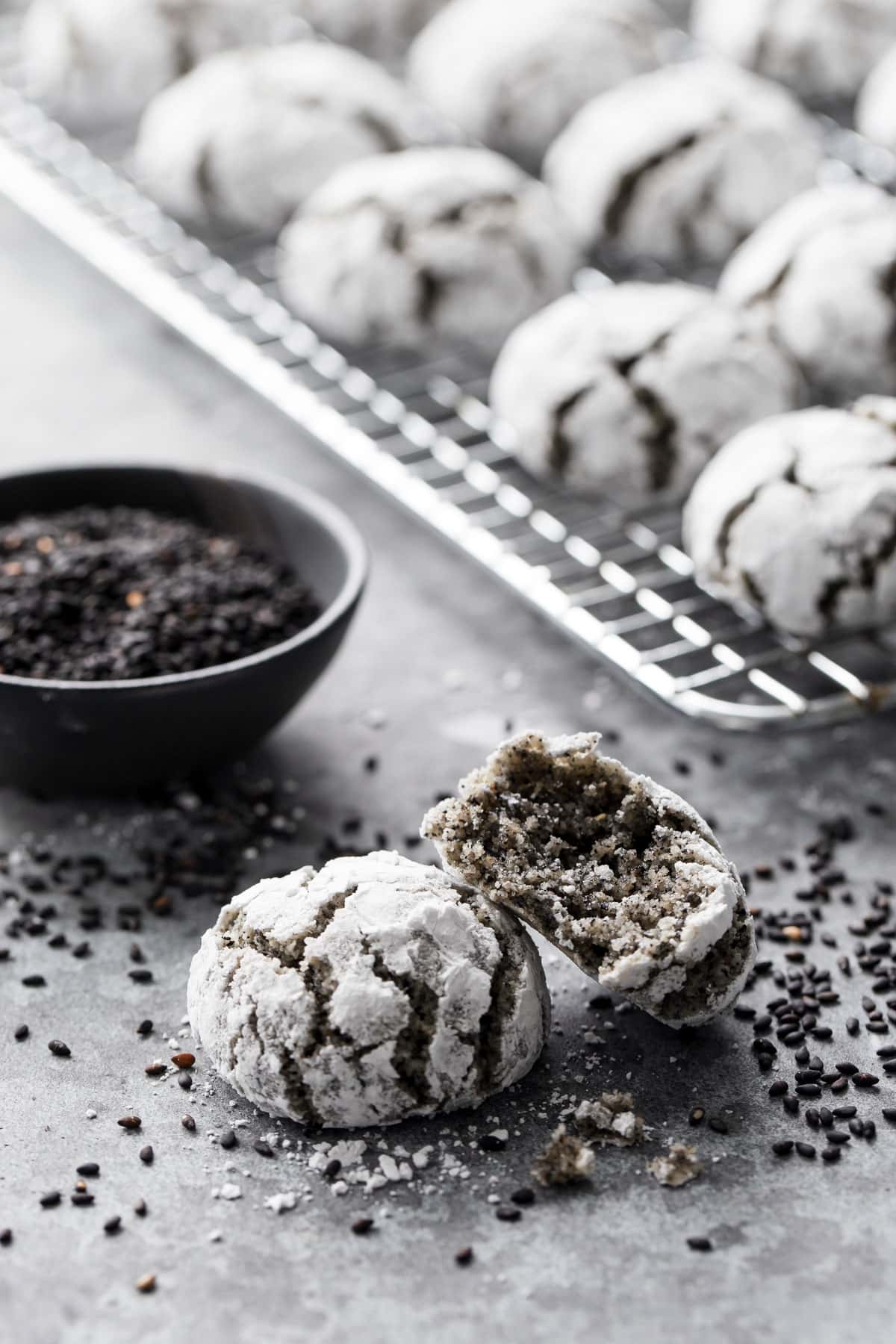 Two Black Sesame Almond Flour Amaretti Cookies, one with a bite out of it to show the interior texture, with a bowl of black sesame seeds and a cooling rack with more cookies in the background.