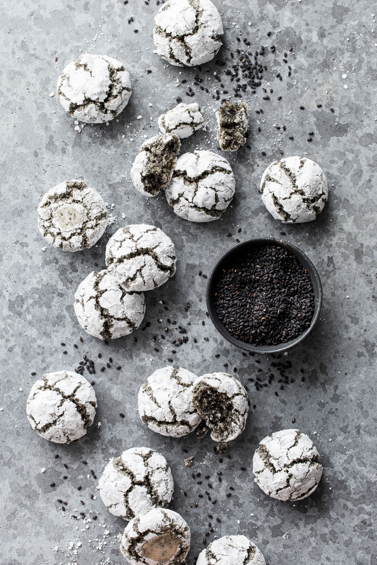 Overhead Black Sesame Amaretti Cookies scattered messily on a gray background with a bowl of black sesame seeds.
