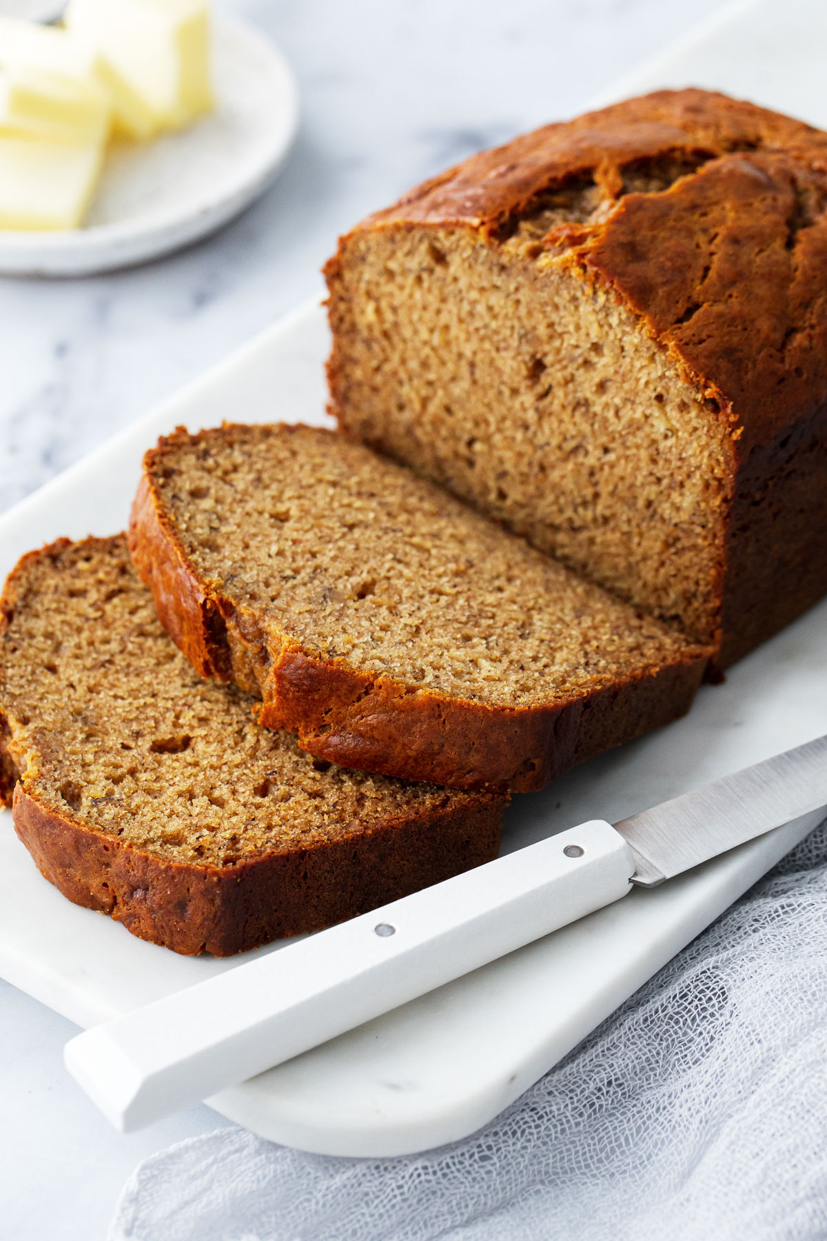 Loaf of Brown Butter Banana Bread on a marble platter, two cut slices laying down to show the interior texture, knife in foreground and dish of butter in the background.